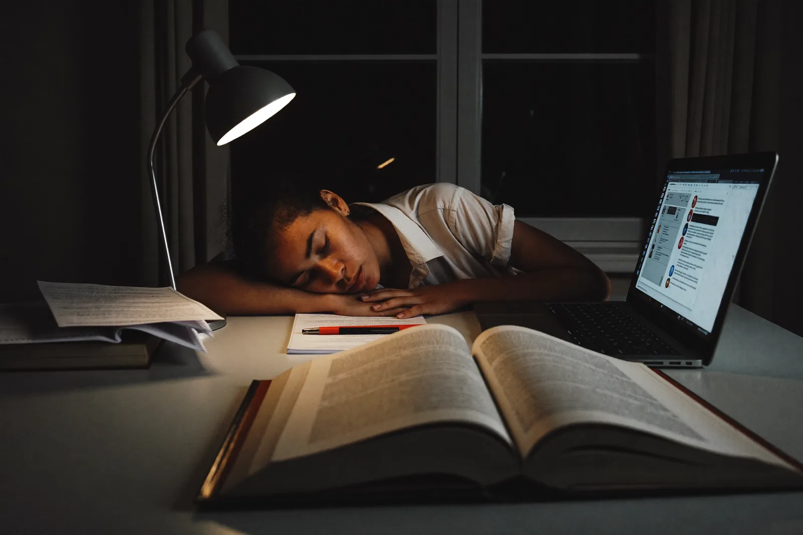 Estudiante dormida sobre sus apuntes de madrugada, junto a una lámpara y un portátil encendido